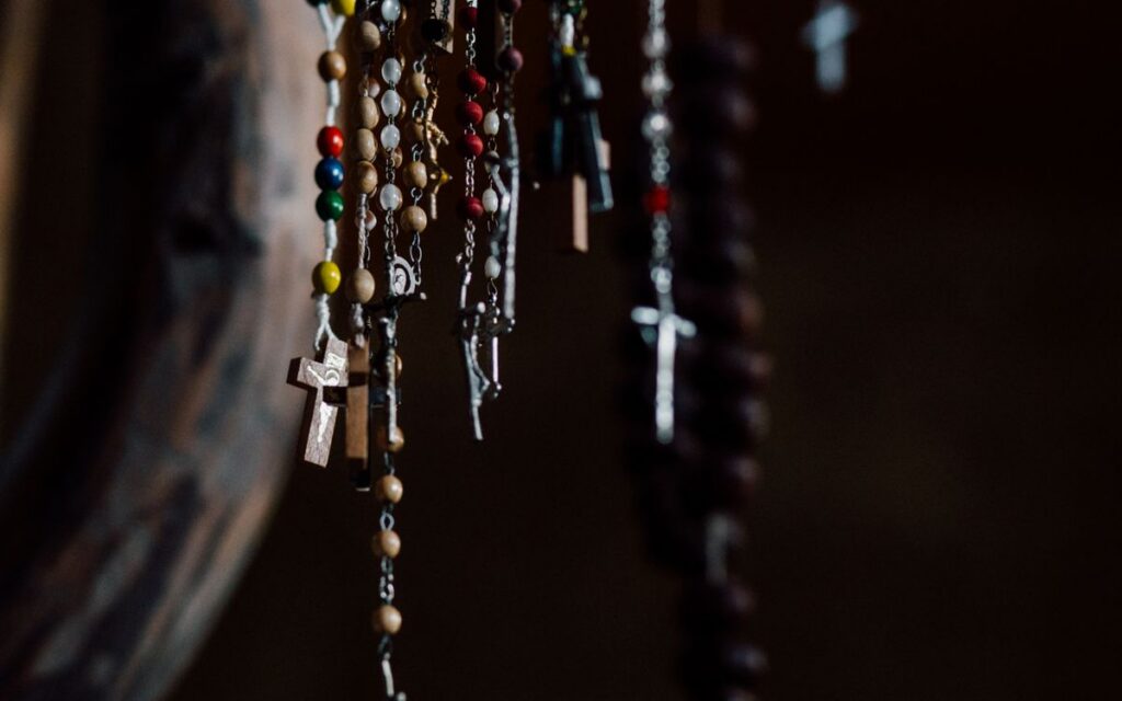 A collection of colorful rosary beads with crosses hanging in a dark indoor setting.