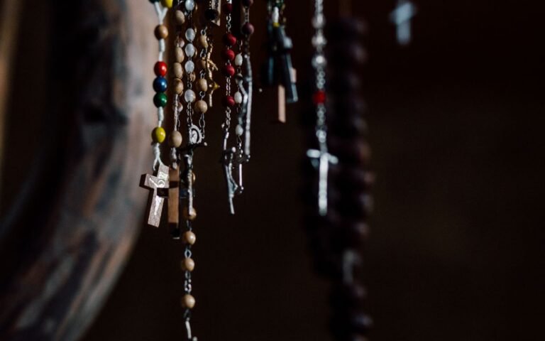A collection of colorful rosary beads with crosses hanging in a dark indoor setting.