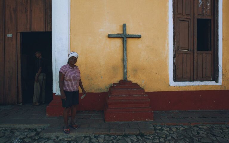 African woman in casual attire standing next to a cross on a rustic building wall.
