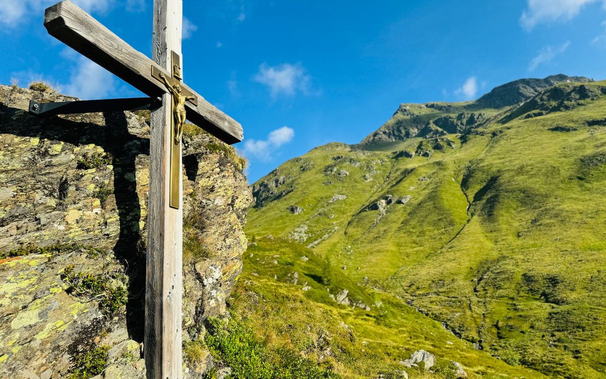 A wooden cross stands on a rocky hill with a mountain backdrop under a clear blue sky.