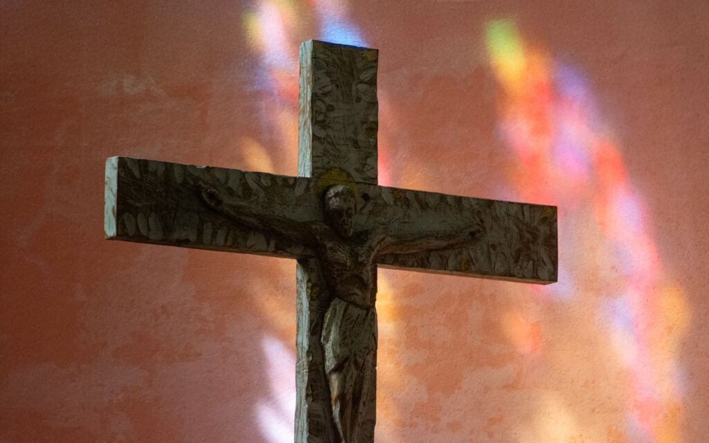 A stone crucifix illuminated by colorful stained glass reflection on an orange wall.