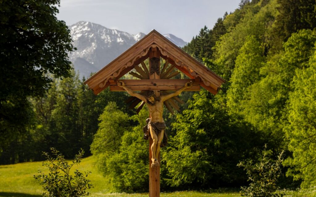 Wooden crucifix set against a lush green forest and distant snowy mountains.