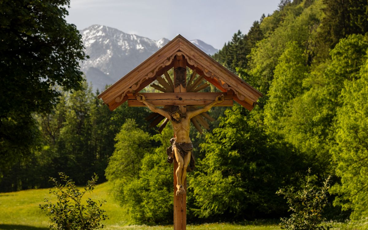 Wooden crucifix set against a lush green forest and distant snowy mountains.