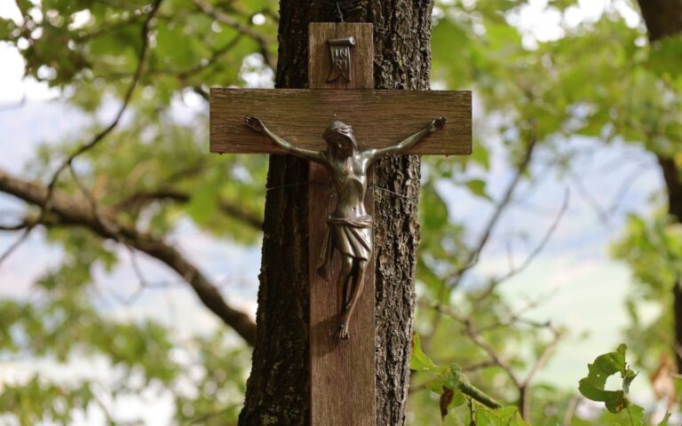 A bronze crucifix on a wooden cross mounted on a tree amidst lush greenery.