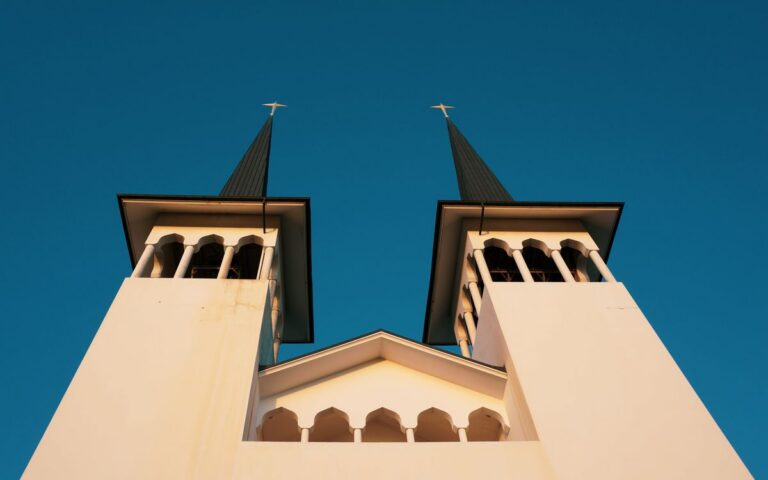 From below of geometric facade of aged church with crosses on sharp roof and arched bell towers against cloudless blue s