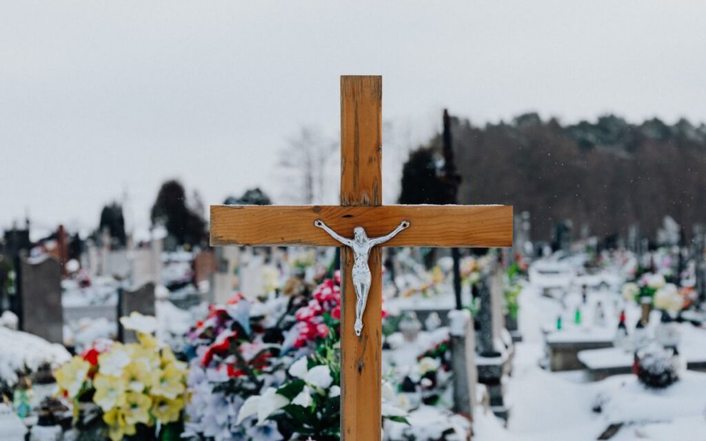 A wooden crucifix stands in a snow-covered cemetery, surrounded by floral tributes.