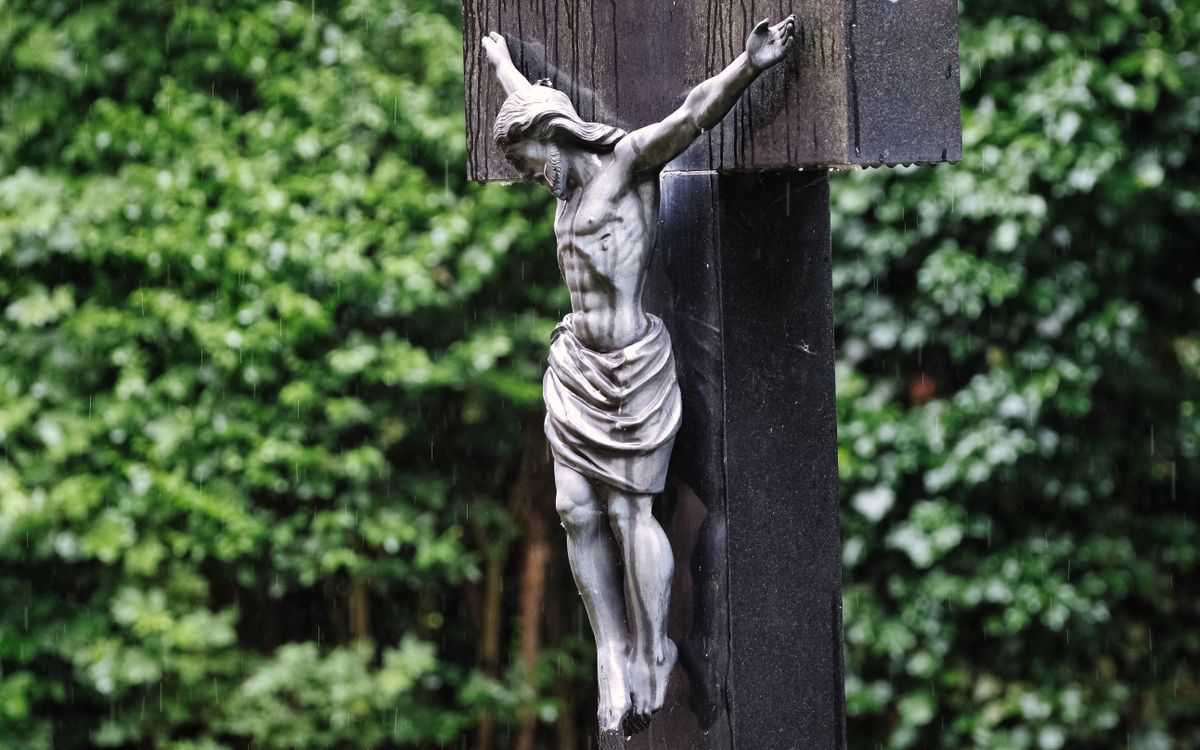A close-up of a wooden crucifix depicting Jesus, set against a green garden background under rain.