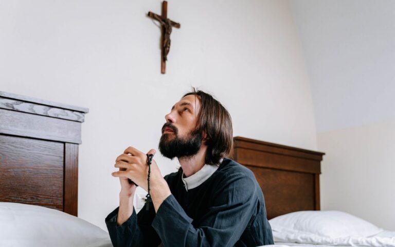 A man kneeling in prayer with a rosary before a crucifix in a cozy bedroom setting.