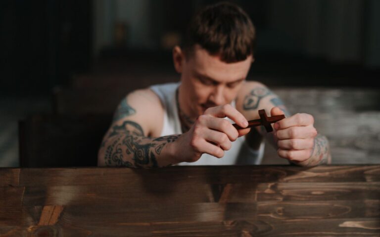 Tattooed man praying with a wooden crucifix inside a dimly lit church, symbolizing faith and reflection.