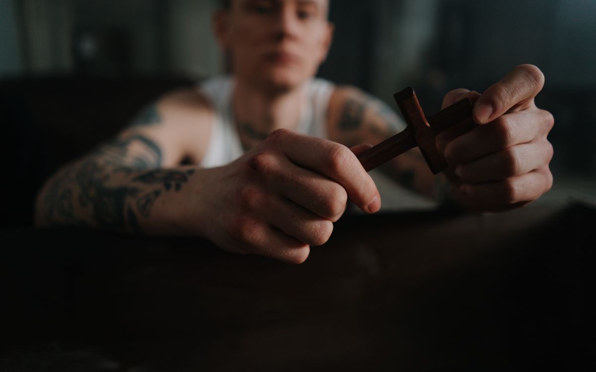 A tattooed man holding a wooden cross, symbolizing faith and contemplation indoors.