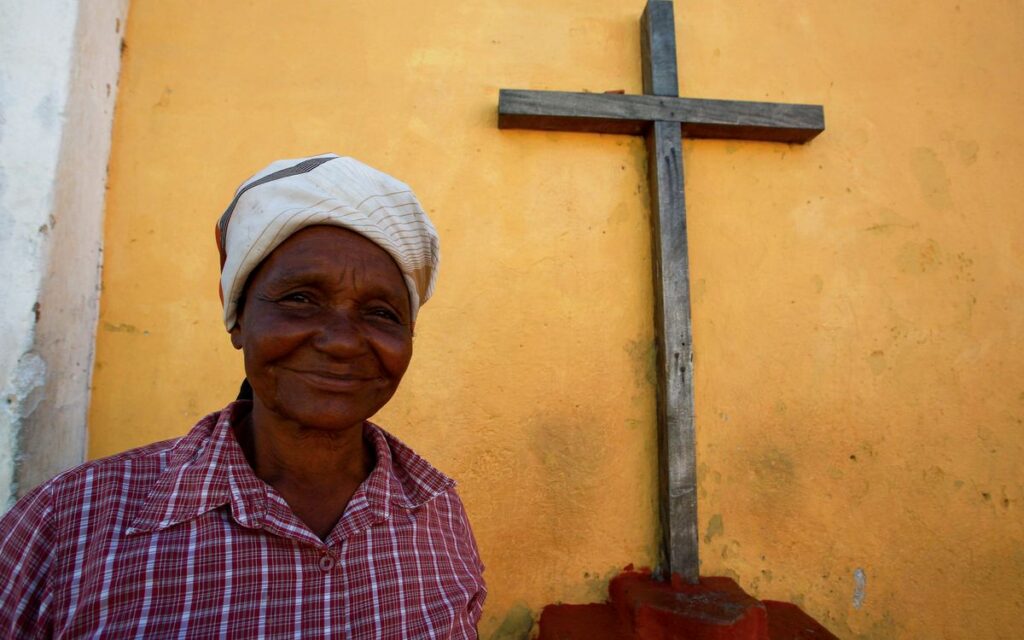 Elderly woman in headscarf smiling beside a wooden cross and yellow wall.