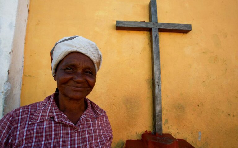 Elderly woman in headscarf smiling beside a wooden cross and yellow wall.