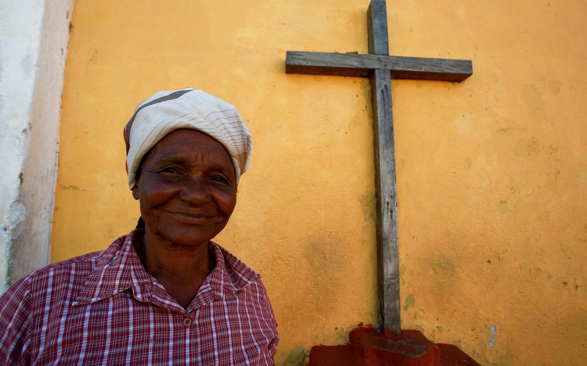 Elderly woman in headscarf smiling beside a wooden cross and yellow wall.