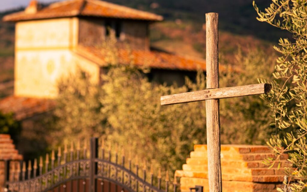 A wooden cross stands in a peaceful rural setting with a rustic building in the background.
