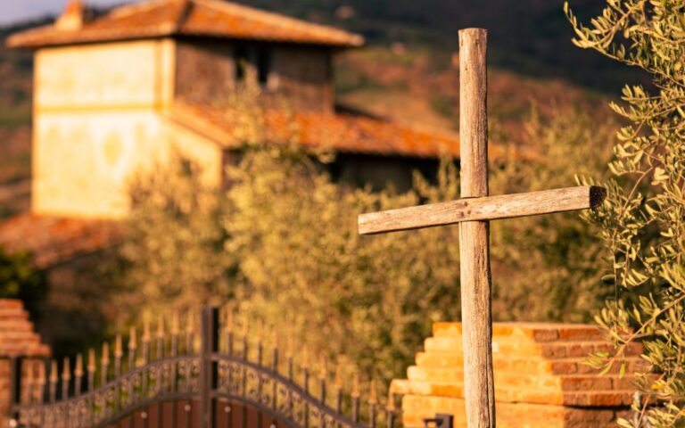 A wooden cross stands in a peaceful rural setting with a rustic building in the background.
