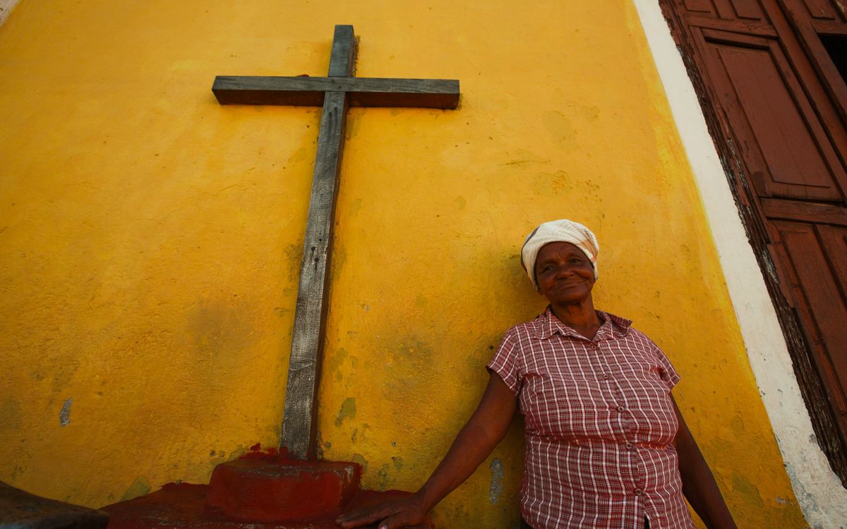 An elderly woman in a plaid shirt stands by a large wooden cross against a vibrant yellow wall.