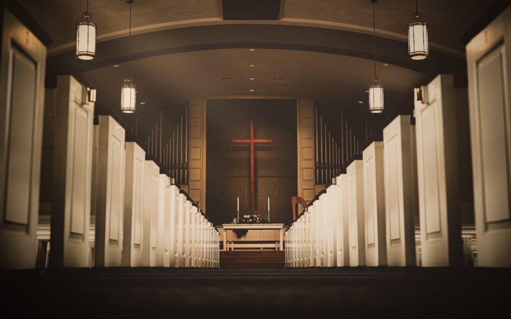 A beautiful church interior with a central aisle leading to a cross and altar, illuminated by hanging lights.