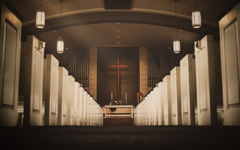 A beautiful church interior with a central aisle leading to a cross and altar, illuminated by hanging lights.