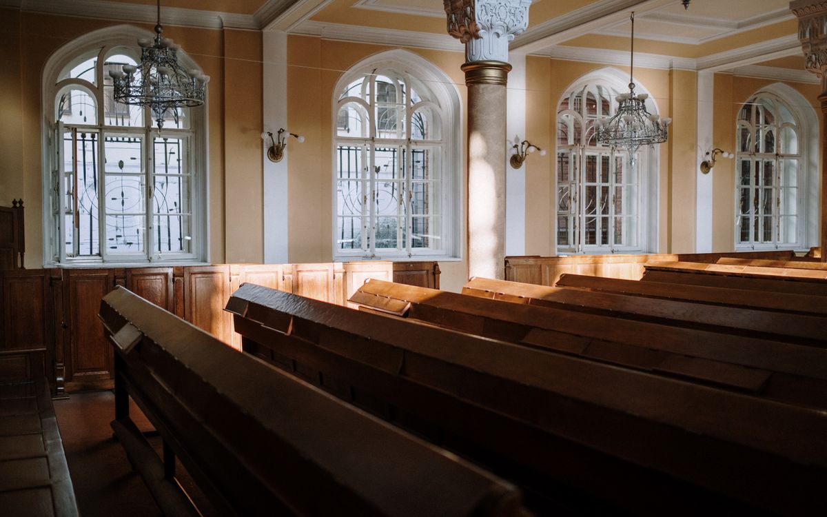 Sunlit church interior with arched windows, wooden pews, and chandeliers.