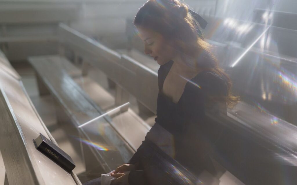A woman sits alone in pews, lost in thought, surrounded by ethereal light.