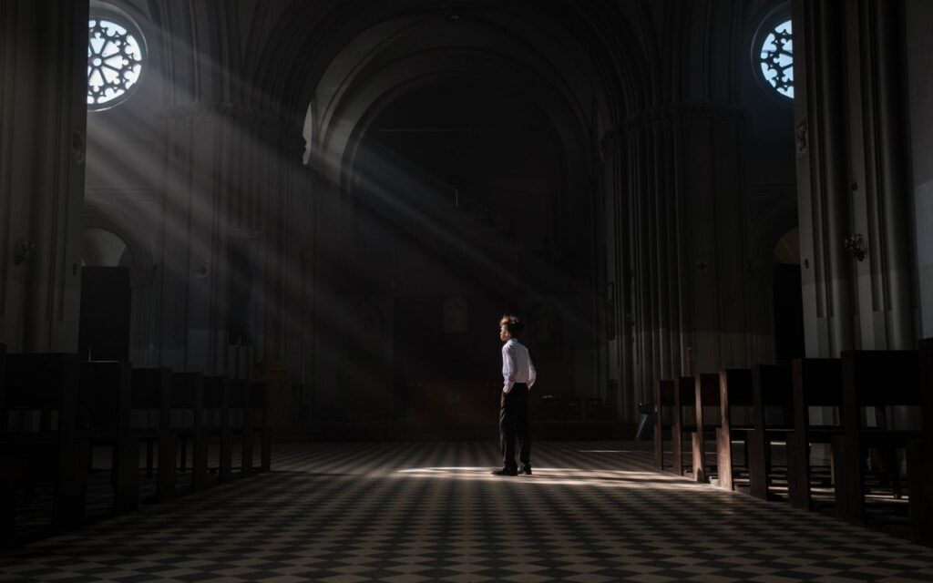 A young boy stands alone in the sunlit interior of a church, casting dramatic shadows.