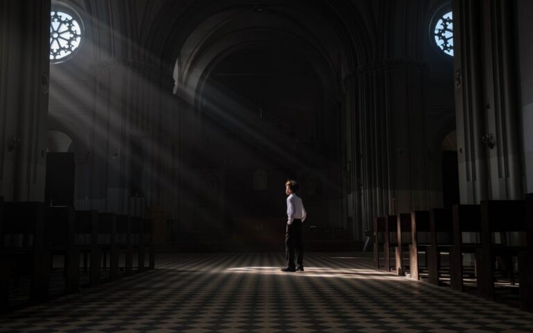 A young boy stands alone in the sunlit interior of a church, casting dramatic shadows.