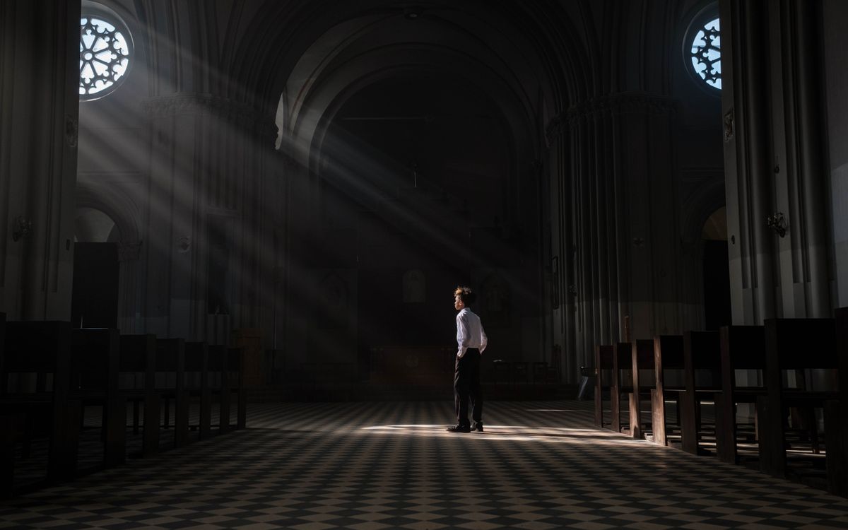 A young boy stands alone in the sunlit interior of a church, casting dramatic shadows.