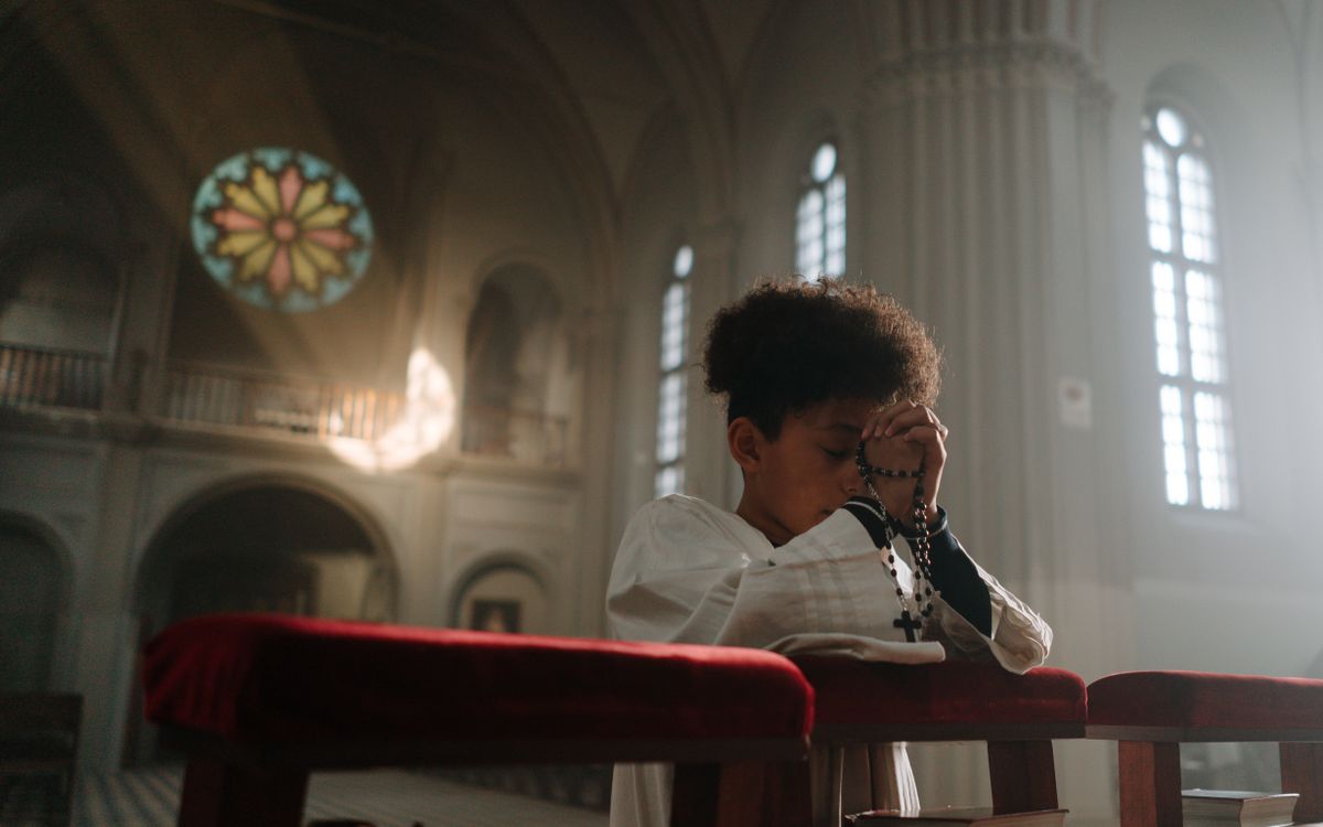 Child praying with rosary in a serene cathedral setting, sunlight through stained glass.
