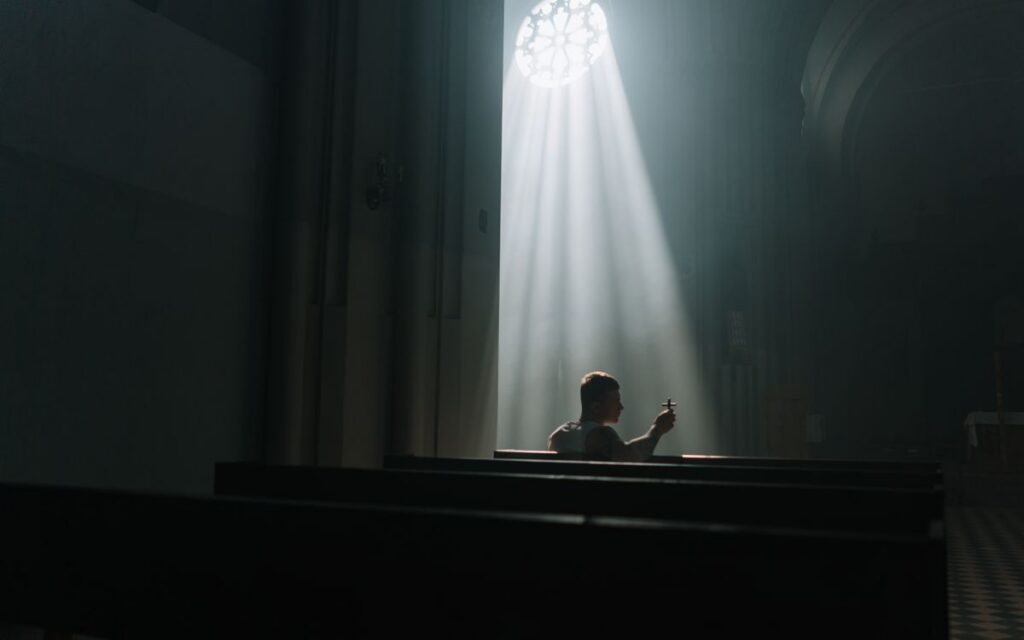 A person sits praying inside a church, illuminated by rays of sunlight through a window.