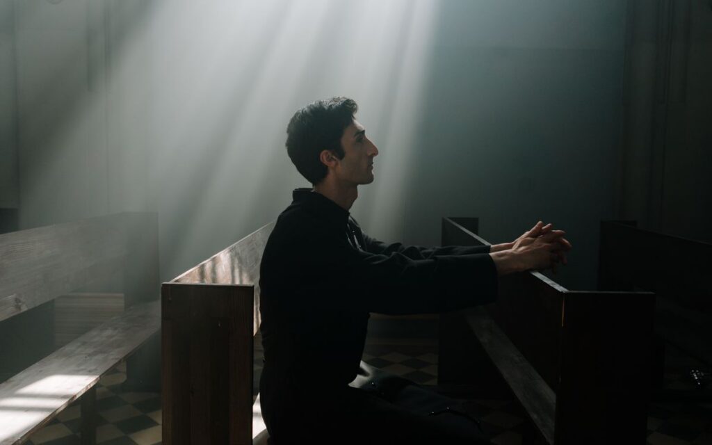 A man sits praying in sunlight inside a dramatic church setting.