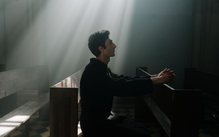 A man sits praying in sunlight inside a dramatic church setting.