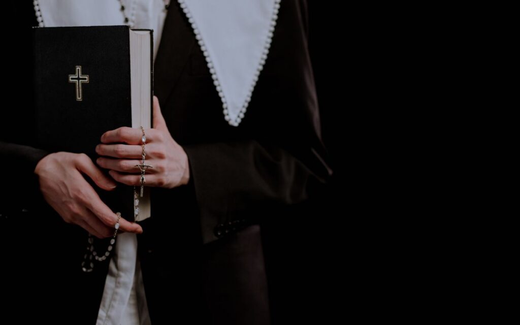 A nun holds a Bible and rosary beads against a dark background, symbolizing faith.
