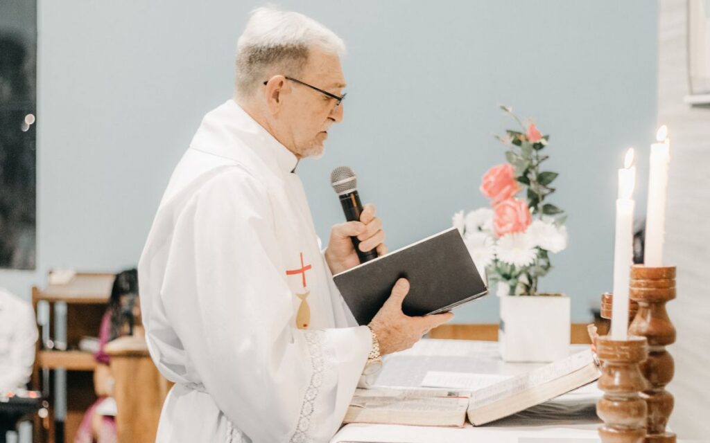 Side view of a Catholic priest in vestments, holding a microphone and book, conducting a ceremony.