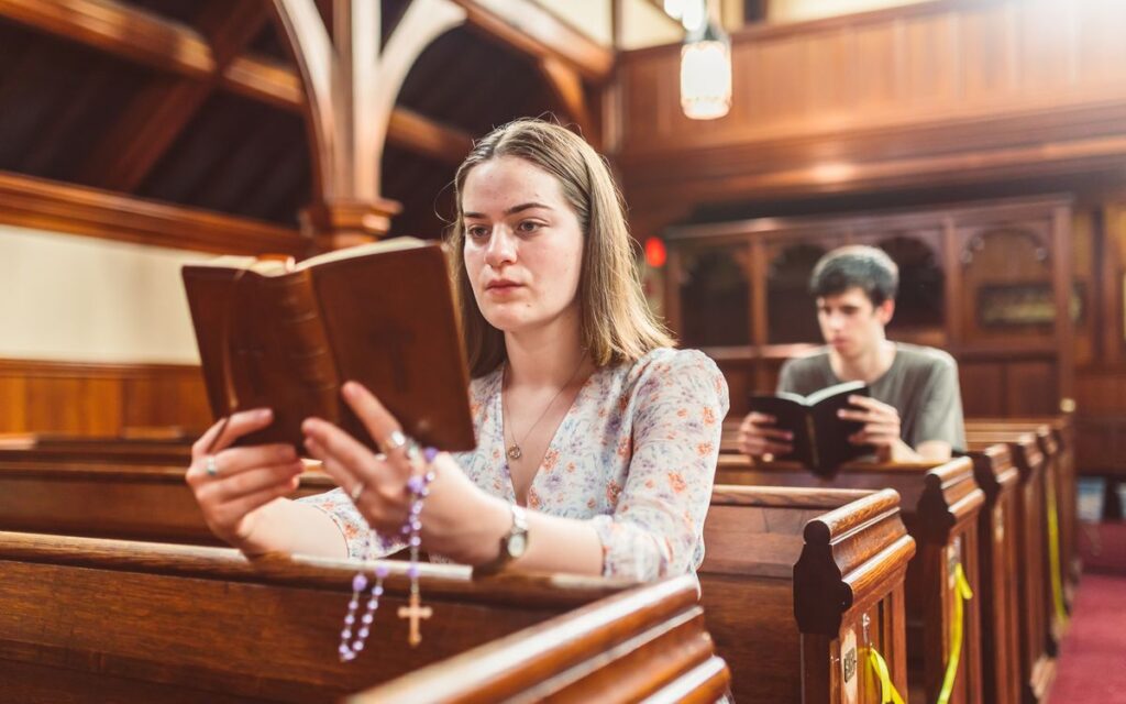 A woman reads a Bible and prays with rosary beads in a church pew.