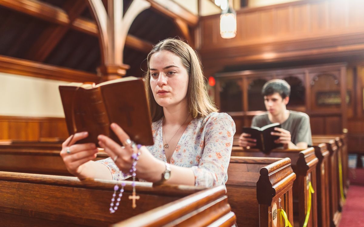 A woman reads a Bible and prays with rosary beads in a church pew.