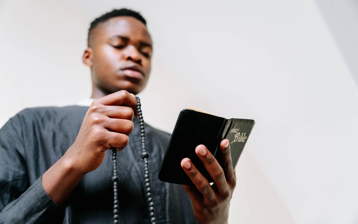 Focused young man holding a Bible and rosary beads in prayer at home.