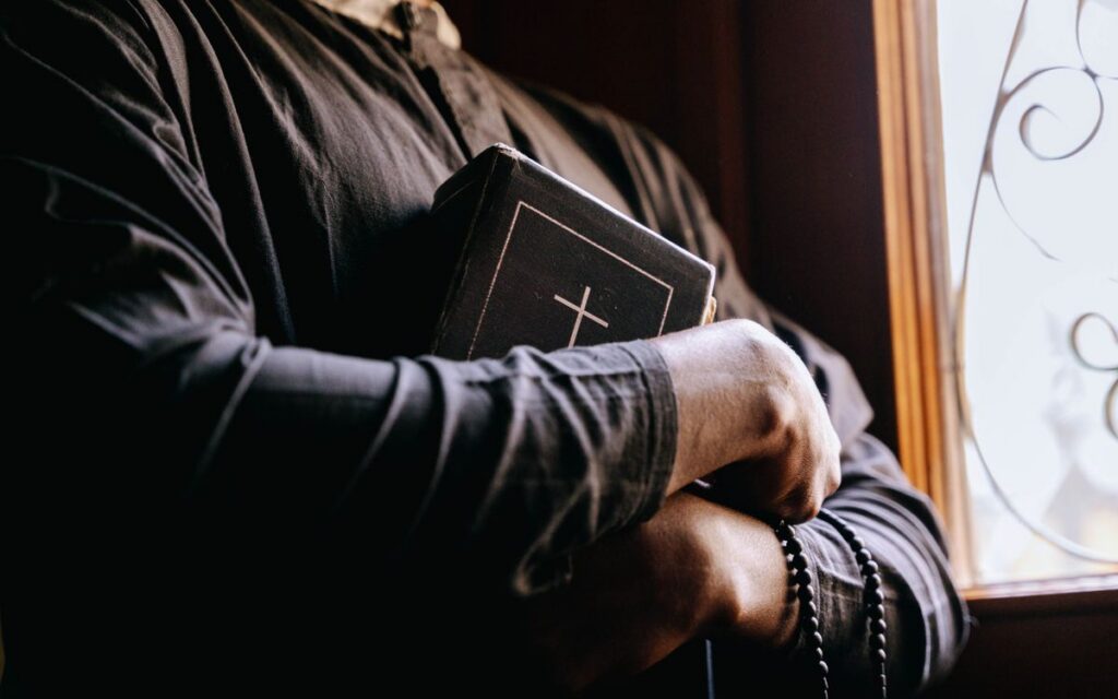 A priest holding a Bible by a sunlit window, symbolizing faith and spirituality.